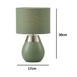 A table lamp with a sage green fabric drum shade and a matching matte green teardrop-shaped base with a brushed silver metal collar.